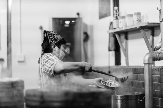 a chef wearing a mask and cap uses a ladle to serve noodles from a noodle boiler in a commercial kitchen